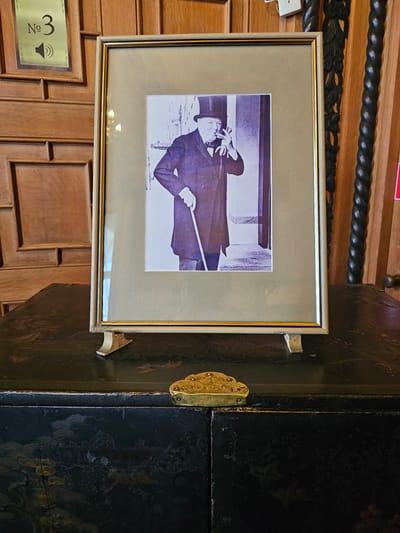 The image shows a framed black and white photograph mounted on a wooden wall, possibly inside a building. The photograph depicts a man in a dark coat and hat, holding a baton or pointer. The photograph is framed with a gold and tan frame. To the right of the framed picture is a dark, glossy cabinet or door, with decorative gold hinges and escutcheon. Below the framed picture, on the wall, there is a plaque that reads "No 3" next to a speaker icon. The wall and objects appear to be located in Алупкинский городской совет, Ukraine, based on geographical indications. The overall environment suggests a historical or formal setting.