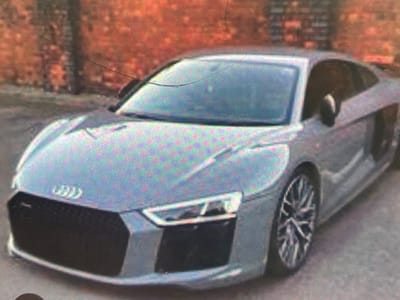 An Audi R8 sports car is parked on a paved surface, possibly a street or driveway. The car is light gray and appears to be in good condition. The background consists of a brick wall, which is out of focus. The overall lighting suggests it is daytime. The car's features, such as its distinctive grille, headlights, and alloy wheels, are visible. There are no people in the image. The scene is located in Paczkowo, Poland.