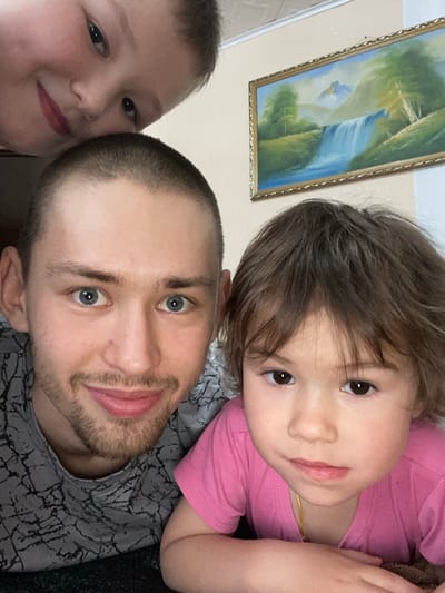 A selfie features three people and a framed painting. The painting, hanging on a wall, depicts a waterfall in a landscape. The selfie includes a man with a shaved head, light blue eyes, and a short beard; a young child with brown hair and brown eyes wearing a pink shirt; and another child with light skin and brown hair. The man wears a gray shirt with a black and white crackle pattern. The location and specific details are insufficient to confirm the picture was taken in Bely Klyuch, Russia.