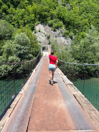 A woman wearing a red t-shirt and light blue shorts walks across a suspension bridge over a clear turquoise river. She has her right arm raised, with her hand open, as if waving. The bridge has metal plating and wooden planks for its surface, with a metal mesh fence along the sides. Lush green trees and a rocky cliff face form the background. The scene appears to be in Moračko Trebaljevo, Montenegro, known for its scenic natural landscapes and rivers.