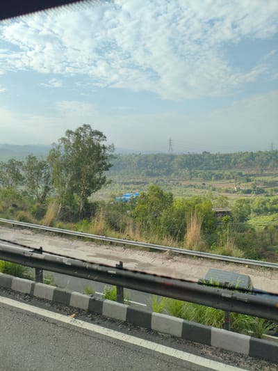 The image shows a view from a road overlooking a valley in Bilaspur, India. The foreground features a road with a concrete barrier and guardrail. Beyond the road, there is a lush green valley with rolling hills, dotted with trees and some scattered buildings. The sky is partly cloudy with a hazy atmosphere. Power lines and transmission towers are visible in the distance, indicating the presence of infrastructure. The overall scene suggests a scenic, rural or semi-rural environment.