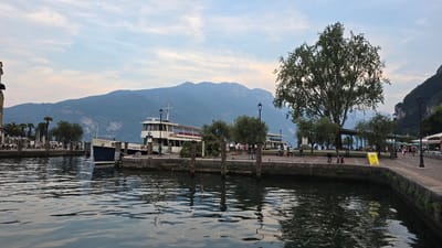 The image depicts a scenic view of the Riva del Garda harbor in Italy. In the foreground, rippling water reflects the overcast sky and surrounding elements. A docked ferry boat, labeled "TRENTO", is prominently featured, its white and blue hull contrasting with the darker water. Behind the ferry, a stone promenade lines the harbor, with people strolling and sitting at outdoor cafes. Several lampposts and trees, including a large, mature tree on the right, dot the promenade. In the background, rolling mountains rise against a soft blue sky with scattered clouds. The overall atmosphere is calm and leisurely, suggesting a pleasant day at the lake.