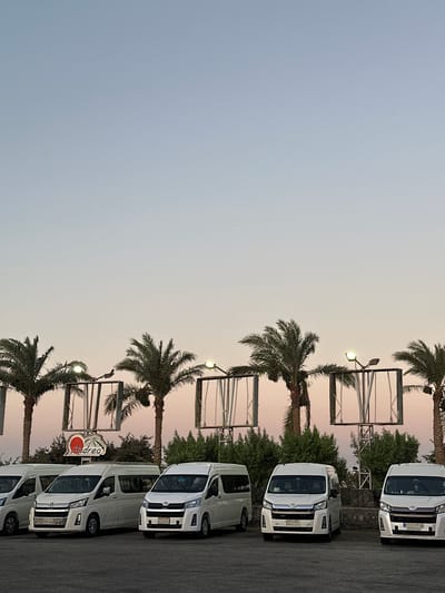 The image depicts a parking lot with several white minivans lined up. Behind the vehicles, a row of palm trees stands tall against a pale sky, suggesting a warm climate. In the background, there are structures that appear to be large outdoor billboards or display screens, some of which are framed by metal scaffolding. A sign with a red heart and the word "Andrea" is visible above one of the minivans. The scene appears to be set in the city of Gharib Cape, Egypt, as indicated by the presence of palm trees and the overall atmosphere. The lighting suggests either early morning or late evening. There are no people clearly visible in the image.