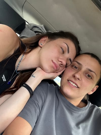 A close-up selfie shows two young women inside what appears to be a car in Dragoman, Bulgaria, during daytime. The woman on the left, with closed eyes, has long dark hair, freckles, a septum piercing, and multiple ear piercings. She rests her left hand on her jawline, revealing a small tattoo on her wrist that reads "have". She wears a black tank top with a white patch visible on her chest, bearing the text "PAUSE JEANS EST.1999", and a beaded necklace with a prominent blue pendant.
The woman on the right, with dark hair pulled back, has her eyes open and is looking towards the camera with a gentle smile, showing her white teeth. She is wearing a grey crew-neck t-shirt and has an ear piercing. The two are positioned intimately, with the woman on the left leaning into the other. The car's light-colored headliner and parts of the seat are visible in the background. The overall atmosphere suggests closeness and relaxation.