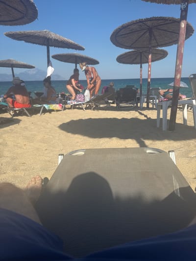 A first-person perspective shot of a beach in Toroni Municipal Unit, Greece, shows a sun lounger in the foreground with a person's legs and feet resting on it. In the midground, beach umbrellas provide shade for several people sitting on loungers. The sea is visible in the background, with mountains in the far distance. The sand is light brown and the sky is clear blue. The scene suggests a relaxed atmosphere on a sunny day.