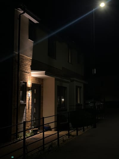 The image captures a nighttime view of a two-story residential building located in Stevenage, United Kingdom. The structure features a ground floor section with a brick facade on the left, transitioning to a light-colored rendered upper story. A dark, vertically paneled front door is visible, marked with the number "13" above an operational exterior wall light on its left. To the left of the door, a small window is present on the ground floor, and another window is on the first floor. A black downspout runs along the far-left edge of the building. Two blue potted plants are positioned on the paved pathway near the front door, which is accessible via a ramp-like section bordered by black metal railings.
The building extends to the right with a consistent modern architectural style, showcasing light-colored rendered sections, a prominent ground-floor column, and multiple windows reflecting internal light sources. The scene is illuminated by a bright, yellow-white street lamp on a tall pole located to the right, casting a distinct diagonal light flare across the upper frame. The foreground pathway, bordered by the railings, recedes into darkness on the right. No individuals, vehicles, or overt activities are present.
