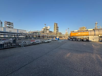 An outdoor, wide-angle photograph captures an industrial complex in Hook of Holland, Netherlands, under a clear, light blue sky. In the foreground, dark gray asphalt pavement exhibits minor cracks. Running horizontally across the mid-ground, a gray metal barrier system separates the paved area from industrial infrastructure. Behind the barrier, a series of large, silver-colored pipes are supported by an elevated metal framework, extending leftward towards a white, flat-roofed industrial building.

To the right, a large semi-trailer tanker truck is parked on the asphalt. The truck features a silver tank with a prominent orange band displaying "MOURIK" in white lettering. The tank unit is supported by multiple axles.

In the background and mid-ground, a dense arrangement of industrial structures dominates the scene. These include towering distillation columns, complex piping networks, and scaffolding characteristic of a chemical or refinery plant. A plume of white smoke or steam emanates from a stack or vent in the central-right portion of the background. Various smaller objects are visible between the barrier and the main industrial structures, including several blue plastic drums and a light blue portable toilet. A distant red and white striped structure is also visible towards the right side of the industrial complex. No individuals or active interactions are depicted.
