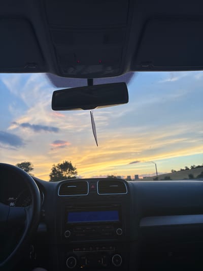 The image shows the interior of a car facing forward, with a view of the sky through the windshield. The sky is a mix of blue and golden hues, indicating it is either sunrise or sunset. Wispy clouds are scattered across the sky, with some appearing pinkish. Trees are visible in the distance on the right side of the frame. The car's dashboard is visible, including the rearview mirror hanging from the windshield, air vents, and a car stereo system with a blue display. The steering wheel is also partially visible in the lower right corner. There is a small, dark object attached to the rearview mirror. The time on the car's display reads "20:01" and the number "86" is visible on the dashboard. The setting appears to be a road trip in a rural or semi-rural area, given the presence of trees and the open sky. The overall mood is serene, likely due to the beautiful sky at either dawn or dusk. The location is Balanovo, Bulgaria.