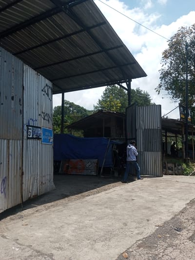 The image depicts an outdoor scene in what appears to be a roadside setting.
**Environment:** The ground is concrete, possibly a road or pathway. There are structures made of corrugated metal sheets forming walls and roofs, creating a sheltered area. Some structures are open, revealing what seems to be a gathering place or workshop. Vegetation, including trees, is visible in the background.
**Subjects/Objects:**
* A person is standing near one of the corrugated metal structures. They are wearing a light-colored hoodie and blue jeans.
* Signage is present on one of the metal walls, with text that includes "BIS 6 CIANJUR" and other words possibly in Indonesian.
* A blue tarp is partially covering something within one of the structures.
**Actions/Interactions:** The person is standing and appears to be either looking at something or talking on a phone.
**Location:** While the prompt mentions Kioumaknao 2, Laos, there's no definitive visual evidence within the image to confirm this precise location. Based on the overall appearance, it could be any roadside area in Southeast Asia or a similar tropical region.