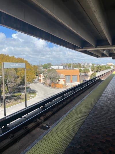 The image provides an elevated view from the platform of the South Miami Metrorail station in South Miami, United States of America. Overhead, large grey concrete structural beams frame the upper portion of the scene. The platform, paved with dark brick-patterned tiles, features a prominent yellow tactile warning strip adjacent to the train tracks. A set of train tracks, including a third rail, runs diagonally from the lower left to the upper right. To the left of the tracks, a white, rectangular station sign on a metal pole clearly displays the text "South Miami." Below the elevated tracks, an asphalt road curves through a vegetated area with several deciduous trees, many appearing bare or with sparse yellow-orange foliage. In the midground, an orange-colored building with two large grey roll-up doors and an attached smaller structure is visible, with red safety cones placed near its entrance. The background consists of a mix of urban architecture, including multi-story residential or commercial buildings, interspersed with palm trees and other vegetation. The sky is partly cloudy with patches of blue. No individuals are present, and no discernible actions or interactions are occurring within the frame.