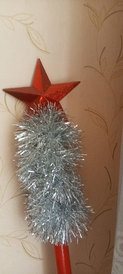 An extreme close-up shot shows the top of a red stick, adorned with silver tinsel and a red star. The background is a beige wallpaper with a subtle, repeating pattern of light brown, stylized leaves. The lighting appears to be indoor, casting soft shadows. No people or actions are depicted, and the scene is static. The image is an indoor shot, likely a home environment, and its location within Zhigulyovsk, Russia, cannot be determined from the visual information provided.