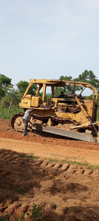 The image captures a daytime scene in what appears to be a rural or land-clearing area, likely Otunade, Nigeria, given the context. The central subject is a large, yellow track-type bulldozer, identified by the visible text "D-8K" on its side, and "CAT." on an interior part, indicating it's a Caterpillar D8K model.

Two men are present with the heavy machinery. One man, wearing a grey t-shirt, dark pants, and a dark cap, is standing beside the bulldozer's tracks, appearing to inspect or work on the lower part of the machine. The other man, dressed in a dark shirt, is seated in the operator's cabin of the bulldozer, suggesting he is either preparing to operate it or is currently operating it.

The bulldozer is situated on reddish-brown, disturbed earth, indicative of recent ground work or a construction site. Clear track marks are visible in the soil around the machine. In the background, there is abundant green foliage and trees under a bright, clear, light blue sky, typical of good weather conditions. The scene implies ongoing land development or maintenance activities. No strong emotions are discernible from the individuals.