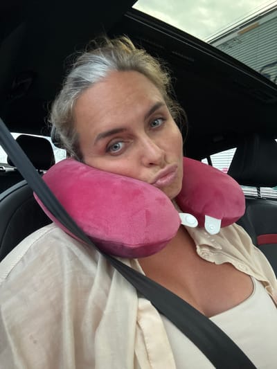 A woman with graying blonde hair is in the passenger seat of a car, wearing a beige button-up shirt and a white tank top. She has a pink travel pillow around her neck and is puckering her lips. The car's sunroof is open, revealing a cloudy sky. The background shows the interior of the car, including the headrest and seatbelt. The image was taken in Ban Nong Bua, Thailand.