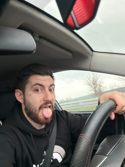 An adult male is situated in the driver's seat of a vehicle, located in Mlada Gvardia, Bulgaria. He possesses dark hair and a beard, and is attired in a black hooded sweatshirt featuring a partially visible white logo. A seatbelt is fastened across his torso. The individual is looking directly towards the viewer with widened eyes and his tongue extended. His right hand is positioned on a black steering wheel, which is fitted with a quilted cover accented with red stitching. The vehicle's interior components visible include a light gray headliner, a partially obscured rearview mirror, a sun visor, and an SRS airbag indicator on the right A-pillar. The exterior environment, visible through the blurred windows, displays a road barrier, deciduous trees devoid of leaves, and an overcast sky, with rain or water droplets evident on the glass surfaces. This suggests the vehicle is in motion under wet weather conditions.