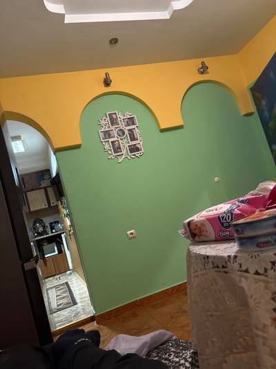 The image captures an interior residential space, likely a living or dining room, connected to a kitchen area. The room features a distinctive wall design with segments of light beige/grey, bright yellow arched trim, and light green paint. A partial blue sky mural adorns a section of the ceiling in the upper left.
Decorative elements include two metallic hooks or fixtures on the yellow trim, and a white, vine-shaped picture frame displaying multiple small photographs on the green wall. A chrome fixture is also present on the beige/grey wall, and a single European-style electrical outlet is visible on the green wall.
In the immediate foreground, a table draped with a white, lacy tablecloth holds a pink package of "Baby Wipes" marked "120 PCS" and featuring an image of two infants. Another object, possibly a book, rests beside the wipes. A hand, clad in a black glove with the partial word "PORT" visible, is seen in the bottom right, resting on a dark surface.
Beyond an archway on the right, a kitchen or utility area is visible, equipped with white appliances, wooden cabinetry, and a patterned, wood-effect tiled floor. A further archway in the background leads to another brightly lit room with a window. The scene suggests a lived-in home environment in Dolna Banya, Bulgaria.