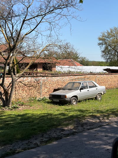 The image shows a silver car parked on a grassy area next to a brick building in Umarevtsi, Bulgaria. The car's license plate reads "OB 3444 AC". A road runs along the edge of the grass. A large, bare tree is visible in the foreground. The sky is blue with some trees in the distance. The building has a red tile roof. The photo was taken in daylight.
