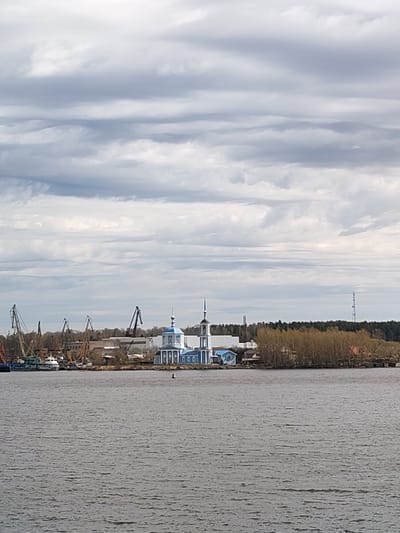 The image shows a body of water with a shoreline in the background. On the shoreline is a blue and white building, likely a church, with a dome and a spire. Adjacent to the church are industrial cranes and buildings. Beyond that is a line of trees and a communications tower. Above, the sky is filled with layered grey and white clouds. Burtsevo, Russia is a possible location given the presence of the Volga River, Russian-Orthodox Church structures, and water crane infrastructure in the area.