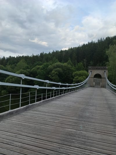 The image shows a wooden suspension bridge in Stádlec, Czechia. The bridge has a weathered wooden deck and white metal railings. In the distance, there is an arched stone tower that anchors the suspension cables. The bridge spans over a body of water, possibly a river, which is dark and reflects the overcast sky. The background is filled with dense green trees, forming a forest that slopes up a hill. The sky is cloudy, with a mix of grey and lighter clouds. There are no people or animals visible in the scene. The lighting suggests it is late afternoon or early morning.