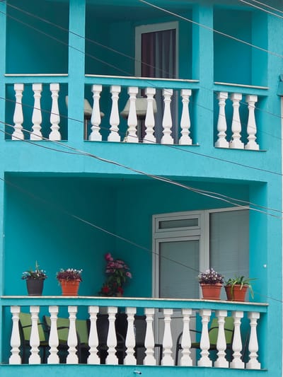 The image shows a close-up of a bright turquoise building with balconies in Ribnovo, Bulgaria. The building has two levels of balconies, each with white balustrades. The upper balcony has two window openings, one with a window with curtains and another empty space. The lower balcony features a window and several potted plants. On the lower balcony, to the left, there are two potted plants with small white flowers in dark pots, and to the right, a larger arrangement of pink and red flowers. In front of the lower balcony railing, there are several green chairs. Power lines are visible crisscrossing the front of the building.