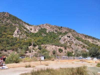 The image depicts a sunny outdoor scene at a natural hot spring in Marikostinovo, Bulgaria. A steep, rocky mountain covered in green foliage dominates the background under a clear blue sky. In the mid-ground, a dirt path leads towards the hot springs, with several people visible. Two women are walking on a wooden platform on the left side of the frame. Further along the path, a woman is sitting on a towel near a pool of water, and another person is partially submerged in the water to the right. Various structures, including a wooden fence, a small shed, and what appear to be changing rooms or small buildings, are scattered throughout the area. Vehicles, including a car and a caravan, are parked in the background. The ground is sandy and dry in most areas, with some grassy patches. The overall impression is one of a recreational area at a natural thermal spring.