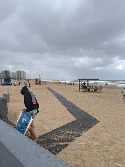 The image captures an overcast day at a beach in Punta del Este, Uruguay. The scene is dominated by a wide sandy shore meeting the ocean, with the horizon largely obscured by a grey, cloudy sky.
In the foreground, a person is seen from behind, leaning over a low concrete barrier. They are wearing a black hooded sweatshirt with a colorful, stylized graphic on the back, and floral-patterned shorts. They are holding a blue cooler, which bears a white logo or symbol, and a part of a white folding chair is visible next to them.
A distinctive dark wooden walkway forms a zigzag path across the light brown sand, leading towards the ocean. Further along the beach, a wooden shelter with benches and tables provides a gathering spot for several people. Numerous other individuals are scattered along the shoreline and near the water's edge, where moderate waves are breaking. A wooden lifeguard stand with a small red/orange flag is also visible in the mid-ground.
In the distance, modern high-rise buildings line the coastline. The weather appears cloudy and muted, suggesting a cool or mild day for beach activities. The colorful graphic on the back of the person's hoodie contains illegible stylized text.