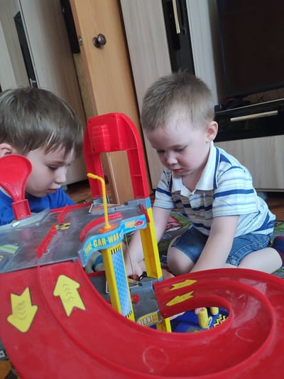 Two young children are playing with a toy car wash on the floor indoors. The child on the left is wearing a blue shirt, while the child on the right is wearing a blue and white striped shirt and denim shorts. The toy car wash is red, grey, and yellow, with features like ramps and a "Car Wax" sign. The location is potentially Malaya Purga, Russia, based on context, but there are no visual clues in the image to confirm this. The children are focused on the toy, suggesting they are engaged in play. The background includes wooden furniture and a television stand.