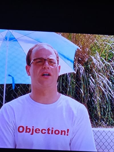 This media file shows a close-up of a person wearing a white t-shirt with red lettering. The person is wearing glasses and appears to be outdoors, with a blue and white umbrella partially visible behind them. Behind the person and umbrella, there is a chain-link fence and some greenery. The t-shirt has the word "Objection!" printed vertically in red letters. The image is tilted, suggesting it might be a candid shot or a still from a video. The lighting suggests it is daytime. The image is from Turka, Ukraine.