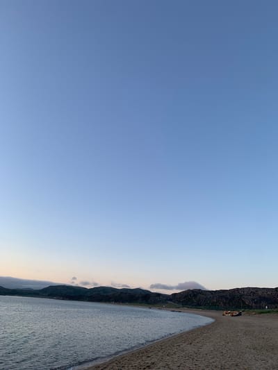 The image shows a wide, sandy beach bordering a calm body of water. In the distance, a chain of dark, rocky hills forms the horizon, with a few scattered structures visible along the shoreline. The sky is a clear, pale blue, transitioning to a lighter hue near the horizon, suggesting either early morning or late evening. On the beach, two yellow pieces of equipment, possibly construction vehicles or boats, are parked. The sand is textured with footprints and ripples from receding waves. The overall scene depicts a serene coastal landscape. The location is the beach in Teriberka, Russia.