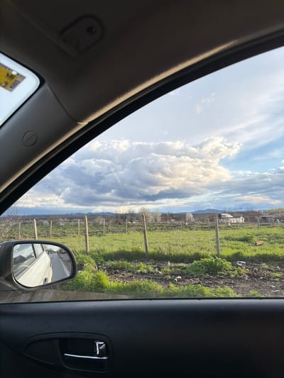 The image is taken from inside a vehicle, looking out the passenger side window. The exterior view shows a rural landscape with a field of green grass and vegetation. A simple wooden fence runs along the edge of the field, separating it from what appears to be a residential area with houses in the distance. The sky is filled with dramatic clouds. The side-view mirror of the vehicle is visible in the lower right corner, reflecting the side of the vehicle itself. The interior of the car is visible in the foreground, including the car door and the upper part of the dashboard. The location in the city of Manastir, Bulgaria is not confirmed.