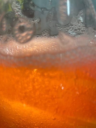 The image displays a close-up view of a beverage in a glass container. The beverage is a vibrant orange color and appears to be carbonated, with numerous small bubbles visible throughout. The top portion of the container shows condensation on the glass, creating a frosted effect, and some dark shapes that could be fruit or ice are visible through the condensation. The lighting creates a shimmering effect on the surface of the liquid. There is no indication of the location within the city of Терволово, Russia, as the image focuses solely on the beverage itself.