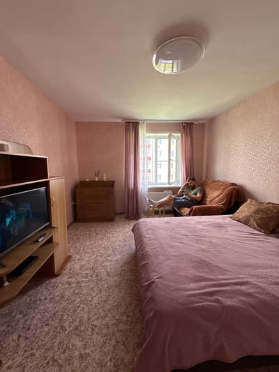 The image shows a bedroom in an apartment in Valday, Russia. A man is sitting on a couch, looking at his phone. The room has pink walls and a patterned carpet. There is a bed, a dresser, and a television. The window is open, and curtains are drawn.