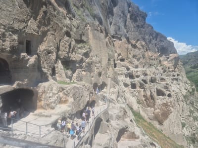 This media file showcases the ancient cave monastery complex of Vardzia, Georgia, carved into a cliff face. The scene depicts a bright, sunny day, likely midday, with a clear blue sky and scattered white clouds. Tourists are actively exploring the site, walking along elevated pathways and stairs that wind through the excavated caves.

The subjects are primarily a group of people, dressed in casual, light-colored clothing suitable for warm weather, suggesting summer. They appear to be engaged in sightseeing, with some pointing and conversing, indicating a leisurely exploration. The pathways are equipped with metal railings for safety, emphasizing the accessibility of the site.

Notable details include the intricate rock-cut architecture of the caves, some of which feature openings that were once dwellings, churches, or refectories. The sheer scale of the cliff and the vast number of carved chambers are evident, highlighting the historical significance and engineering prowess of its creators. The rugged, natural landscape surrounding the monastery, with green slopes and a distant valley, provides a picturesque backdrop.

The overall emotion conveyed is one of awe and discovery, as visitors navigate this remarkable historical and architectural wonder. The sun casts sharp shadows, further defining the textures of the rock and the various levels of the complex. No visible text is present in the image.