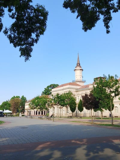 This is a wide shot of a public square in Becej Municipality, Serbia, with a building in the background. The square is paved with grey bricks and features several benches. There are trees along the side of the square, and a large building with a prominent tower is visible. The sky is clear and blue.