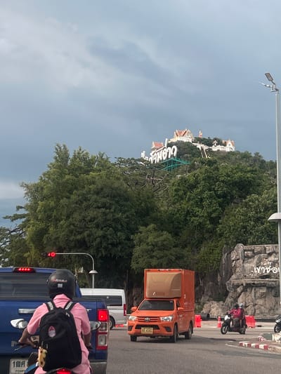 The image captures a street scene in Prachuap Khiri Khan, Thailand. In the foreground, a person wearing a pink shirt and black helmet is riding a motorcycle. They are carrying a black backpack. The motorcycle is behind a blue vehicle. Further in the background, an orange truck is parked on the side of the road. Beyond the road, a lush green hill is covered with trees. A white building with a distinctive architectural style, possibly a temple or a resort, is visible on the hillside. A large sign with Thai script is also present on the hill. The sky is overcast and appears to be either early morning or late afternoon. The overall impression is a typical day in a Thai town with both natural and man-made elements present.