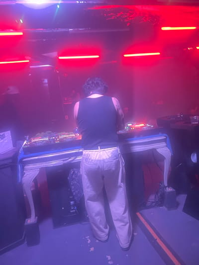 An individual is depicted from the rear, standing in a dimly lit indoor environment, specifically identified as 강남구, South Korea. The person wears a black sleeveless top and light-colored trousers, with dark, wavy hair and visible tattoos on their left arm. They are positioned behind a white, ornately carved console table that serves as a DJ booth, actively engaging with electronic music equipment featuring numerous illuminated controls, buttons, and faders.
The scene is dominated by strong red lighting from horizontal strip lights mounted on the ceiling, creating a pervasive red and purple hue. The floor is dark with scattered white debris. To the left of the main subject, a partial figure wearing a light-colored baseball cap is visible, along with white items on a surface. A dark object, possibly a speaker, and a clear plastic cup containing a beverage are present on the right side of the DJ setup. An orange line is marked on the floor to the right. The overall environment suggests a nightclub or music venue.