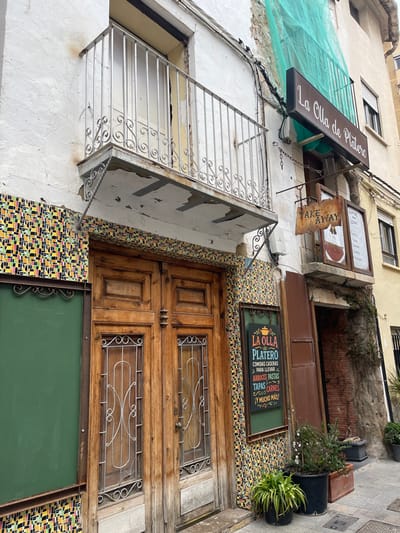 The image displays a street scene in Buñol, Spain, focused on the exterior of a building identified as "La Olla de Platero" restaurant. The ground floor entrance features a double-leaf, dark brown wooden door with glass panels and decorative metal grilles. The facade immediately surrounding the entrance is clad in a mosaic of small, multi-colored rectangular tiles in hues of yellow, red, black, and green. To the right of the door, a vertically oriented black chalkboard sign with a green background displays "LA OLLA de PLATERO" and lists menu items such as "ARROCES, PASTAS, TAPAS, CARNES" in colorful text. A large, blank green board is mounted to the left of the door.
Above the entrance, a white metal balcony with an ornate scrollwork railing is affixed to the building; its underside shows visible wear, including peeling paint and exposed concrete, with rust stains evident on the white stucco wall below. Higher on the facade, a rectangular dark sign with "La Olla de Platero" in white text is positioned above a green awning. Below this, a rustic, irregular-shaped wooden sign proclaiming "TAKE AWAY" hangs by chains. Exposed electrical conduits run along the white stucco wall. To the right, a section of the building's upper wall is partially covered by a light green construction net, and an adjacent cream-colored building with dark-framed windows is visible. Several potted plants are arranged on the light-colored paving stone sidewalk in front of a dark, recessed doorway on the far right. No individuals are present in the depicted scene.