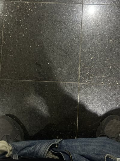 The scene, located in Rafz, Switzerland, depicts a first-person, downward view of an individual's lower body. The subject is wearing blue denim jeans, partially unzipped at the fly, and dark grey athletic shoes with black outsoles. They are standing on an interior floor composed of dark, speckled square tiles, approximately 30x30 cm, separated by lighter colored grout lines forming a grid pattern. Shadows, consistent with a human form, are cast forward from the individual onto the tiled surface. A distinct, bright reflection from an overhead light source is visible on the upper right portion of the polished floor tiles. No other individuals or objects are discernible in the frame.