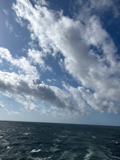 The image depicts a vast expanse of dark, choppy ocean under a bright blue sky filled with scattered white clouds. The horizon line is sharp, separating the sea from the sky. The water is turbulent, with whitecaps indicating movement. In the lower right corner, a white wake trails behind, suggesting a vessel is present, though not visible in the frame. The overall impression is one of an open sea under a dynamic sky. The location is not Żarnowiec, Poland, as the image shows an ocean.