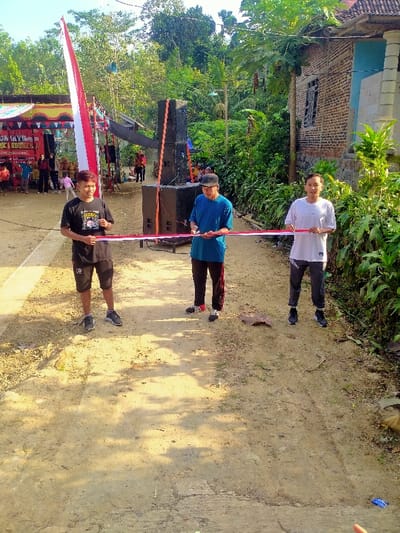 An outdoor scene in Jetis, Indonesia, depicts a ribbon-cutting ceremony on an unpaved dirt path under clear skies. Three males are positioned across the path in the foreground. At the center, an older male, wearing a blue short-sleeved shirt, dark trousers with red stripes, and dark athletic shoes, holds silver scissors in his right hand, poised to cut a red and white ribbon. To his left, a younger male with reddish-brown hair, wearing a black t-shirt, black shorts, and dark athletic shoes, holds the left end of the ribbon with both hands. To his right, another young male, wearing a white short-sleeved t-shirt, dark track pants, and dark athletic shoes, holds the right end of the ribbon with both hands.
In the left mid-ground, a yellow and orange striped tent is visible, decorated with colorful bunting and banners, one of which partially displays "MUNGKIN KITA INDONESIA." Approximately 10-15 individuals, including children, are gathered near this tent, observing the foreground activity. Adjacent to the tent, two large, black sound system speaker stacks are present, secured with orange straps. The background on the right features dense green tropical vegetation, including a visible papaya tree, and the partial facade of a brick house with a tiled roof and a blue-painted section, featuring a dark-framed window. The foreground path is composed of packed dirt with some uneven patches and scattered shadows from overhead foliage.