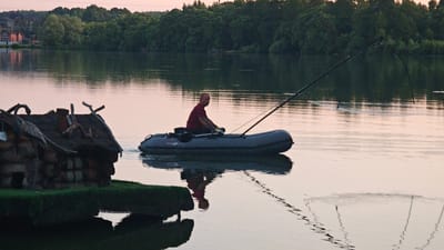 A middle-aged man with a shaved head, wearing a maroon t-shirt and yellow gloves, is fishing from a gray inflatable boat on a calm body of water. He is holding a long fishing rod that is bent, suggesting he may have a catch. The boat has a black oar resting inside. To the left of the boat, on the shore, is a small, rustic wooden structure resembling a cabin, with a thatched roof and decorative bird carvings. The background consists of a dense line of trees and houses in the distance, reflecting on the water's surface. The sky is a soft pinkish hue, indicating either sunrise or sunset. The scene is located in Щапово, Russia.