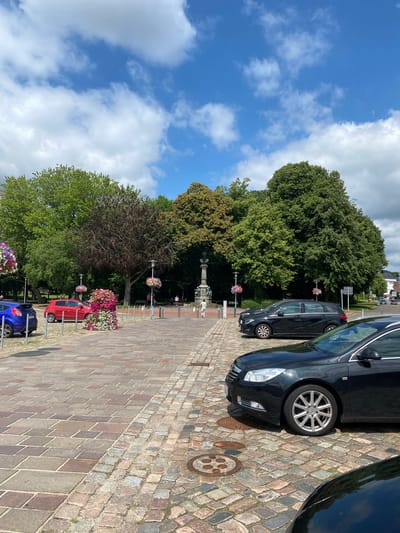 The image depicts a public square in Rendsburg, Germany, likely near the historic town center. The foreground is dominated by a cobblestone pavement. Several cars are parked on the square, including a black sedan in the immediate foreground, another black station wagon behind it, and a red car further back. A blue hatchback is parked to the left. Lush green trees form a dense backdrop, with a statue or monument visible in the mid-ground, surrounded by flower arrangements hanging from lampposts. A person in a light-colored outfit is walking towards the monument. The sky is blue with scattered white clouds.