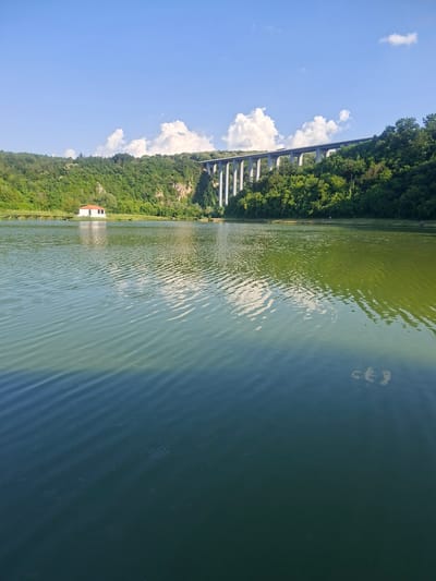 The image depicts a serene landscape with a body of water in the foreground, reflecting the sky and surrounding vegetation. On the left side of the water, a small white building with a red roof is visible, situated near a wooden pier extending into the lake. In the background, a lush green hill covered with dense trees rises, and a large, modern viaduct with multiple concrete pillars spans across the top of the hill. The sky is blue with scattered white clouds. The overall scene suggests a tranquil outdoor setting, possibly a park or a recreational area. The location is identified as Pisanets, Bulgaria.