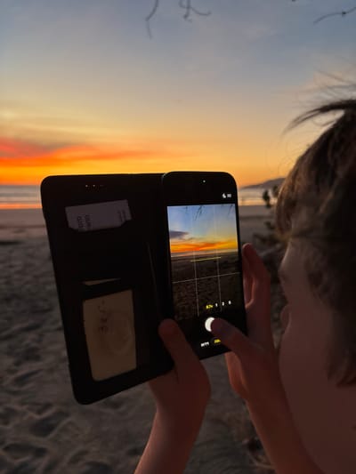 A person is holding a smartphone to take a photograph of a sunset. The phone is in a black case that is open. The background appears to be a beach at Cabo Velas, Costa Rica. The grid lines are enabled on the phone's camera.