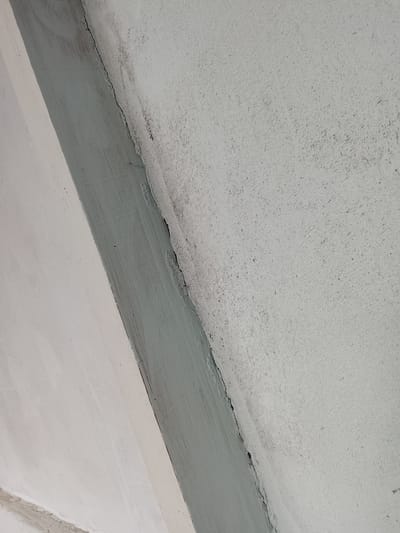 The image shows a close-up, angled view of a textured, light-colored wall with a green-painted strip running diagonally across it. A thin, dark crack is visible along the edge where the painted strip meets the wall. The wall surface is rough and speckled with small dark particles, suggesting it is made of concrete or stucco. There are no people or specific landmarks visible to indicate a precise location within Mehnatabad, Tajikistan. The scene appears to be an interior or exterior wall detail.