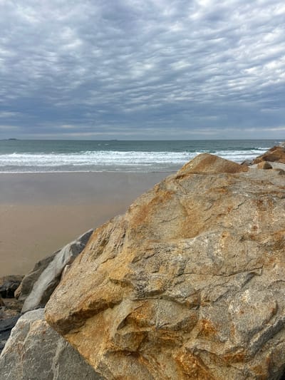 The image depicts a rocky shoreline with the ocean and sky in the background. In the foreground, large rocks, some with a reddish-brown hue and others grayish, are prominent. Beyond the rocks lies a sandy beach with gentle waves approaching the shore. The ocean stretches to the horizon, where it meets a cloudy sky. Distant landmasses or islands are faintly visible on the horizon. The overall scene is one of a natural coastal environment, likely photographed during overcast weather. This scenery is consistent with the coastal areas around Coffs Harbour, Australia, known for its rocky headlands and beaches. There are no people or animals visible in the image.