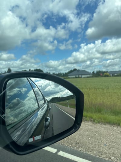 This image captures a view from the passenger side of a car, looking towards the side mirror. The mirror reflects the sky, clouds, and a portion of the car's body. The main scene outside the car, visible on the left, is a sky filled with dramatic, fluffy white clouds against a bright blue background. Below the sky, a green field stretches out, with some trees and buildings visible in the distance, suggesting a rural or semi-rural setting.
The side mirror itself is a prominent object, framed by a black casing. The reflection within the mirror shows a road and the car's interior or body. A notable detail in the reflection is the word "AUTODAMS" printed vertically in red and white letters on what appears to be a sticker on the car's window or bodywork, as seen in the mirror.
The lighting suggests it is daytime, likely mid-morning or afternoon, given the bright sky and the presence of shadows. The weather appears to be fair with scattered clouds. The overall impression is one of travel, possibly on a road trip through a scenic area. The perspective implies the photo was taken by someone in the passenger seat. The location context of Ilūkste, Latvia, places this scene in the Baltic region.