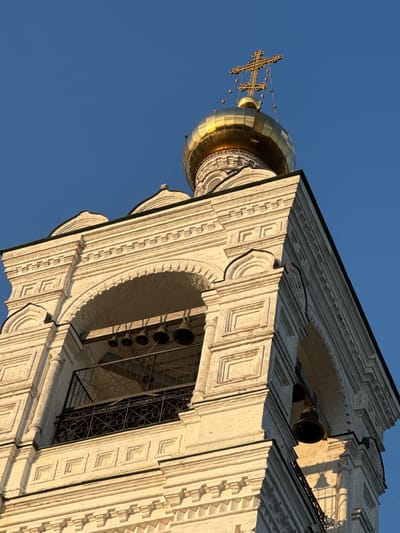 This is a low-angle, close-up, eye-level, outdoor, daytime, medium shot of a white brick bell tower with a golden dome in Zagornovo, Russia. The sky is a clear, deep blue. The bell tower has multiple arched openings containing bells and a decorative metal railing. Intricate brickwork and decorative elements are visible on the tower's facade. A golden dome with a cross adorns the top of the structure. The lighting suggests the sun is low in the sky, casting warm light and shadows on the building. No people are visible in the image.