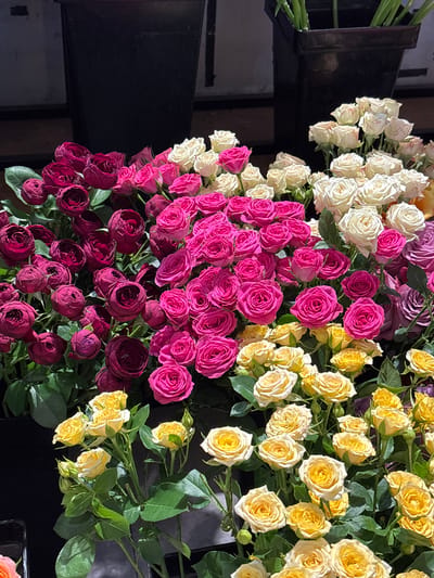 The image displays a close-up, eye-level view of multiple bouquets of roses in various colors, including deep red, hot pink, white, and yellow. The roses are densely packed and arranged in what appears to be a flower shop or market. In the background, dark, tall rectangular containers are visible, suggesting they are being used to hold the flowers or for display. The lighting is somewhat dim, with some highlights on the petals. No people are visible in the frame. The scene is a still life focusing solely on the floral arrangements. There is no information available within the image to suggest a specific location in Shenhe District, China.