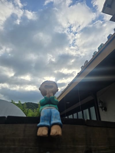 The image depicts a garden figurine of a child with its hands pressed together, positioned on a wooden fence. The figurine is facing upwards towards a cloudy sky. Behind the figurine, a building with a tiled roof and large windows is visible, along with a lamppost mounted on the wall. The background also features greenery and a greenhouse-like structure. The overall scene suggests an outdoor setting in Gumi-si, South Korea.