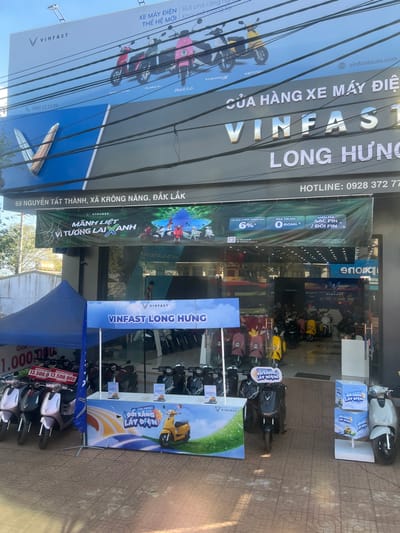 The image displays the exterior of a VinFast electric motorcycle dealership, specifically identified as "VINFAST LONG HƯNG," situated at 69 Nguyen Tat Thanh, Krong Nang, Dak Lak Province, Vietnam. The building features a multi-level facade under clear daylight. The uppermost blue banner advertises "XE MÁY ĐIỆN THẾ HỆ MỚI" (New Generation Electric Motorcycles) and showcases five distinct VinFast electric scooter models: Vento S, Theon S, Feliz S, Klara S, and Evo 200. Below this, a large grey sign prominently displays the VinFast logo and the store name "VINFAST LONG HƯNG," with the full address "69 NGUYEN TAT THANH, XA KRONG NANG, ĐẮK LẮK" and a hotline number "0928 372 777" visible. A green banner beneath advertises promotions, including "MẠNH LIỆT VÌ TƯƠNG LAI XANH" (Strong for a Green Future), a 6% interest rate, 0 down payment, and free battery charging/swapping. Numerous utility lines crisscross the sky above the signage.
On the tiled pavement in the foreground, a promotional setup is active. To the left, a blue pop-up canopy covers several VinFast electric scooters, predominantly in light purple/pink and white, with a price display stating "13.500.000". In the center, a blue and white promotional booth for "VINFAST LONG HƯNG" displays a graphic of a yellow scooter and the slogan "ĐỔI XĂNG LẤY ĐIỆN" (Swap gasoline for electricity), with several brochures placed on its counter. A black VinFast scooter is positioned directly in front of this booth. Additional dark-colored VinFast scooters are parked in rows behind the booth. To the far right, a white VinFast scooter is presented next to a stack of blue promotional boxes.
Through the large glass entrance of the store, numerous VinFast electric scooters of various colors, including yellow, red, black, pink, white, and green, are visibly displayed on a polished interior floor. One male individual is partially visible standing inside the store in the background, near the scooters.